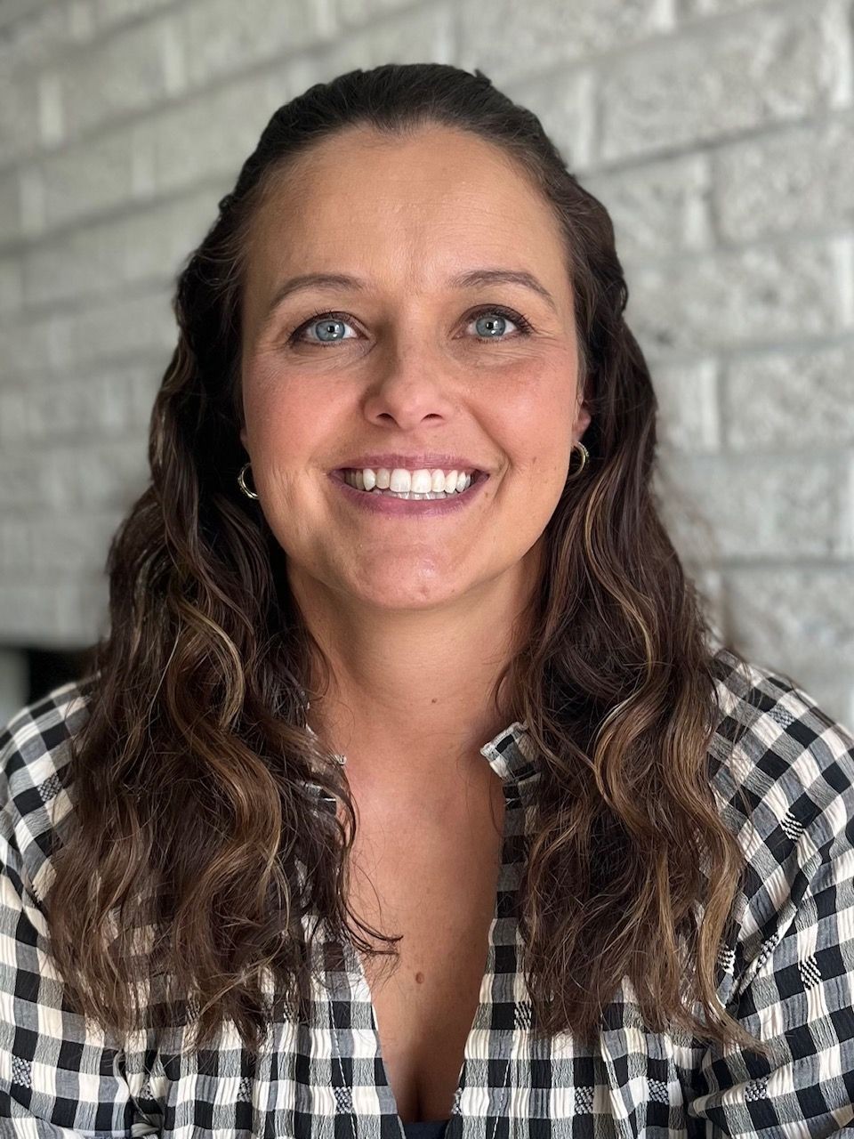 Smiling woman with long wavy hair and checkered shirt against a brick wall background.