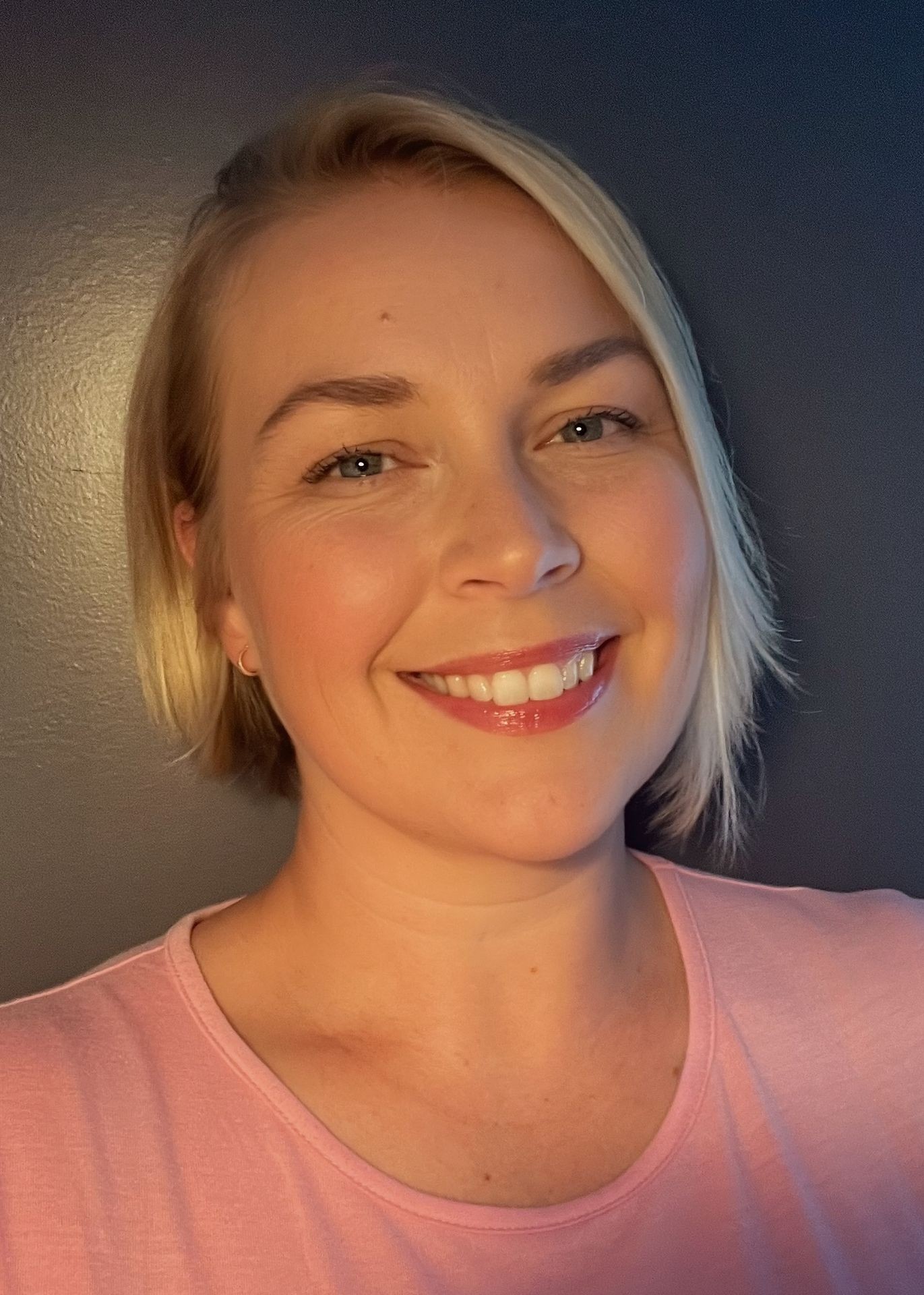 Smiling person with short blonde hair wearing a pink shirt against a dark background.